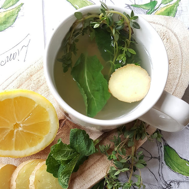 Overhead view of a mug of herbal tea with mint, thyme, and ginger, beside lemon slices